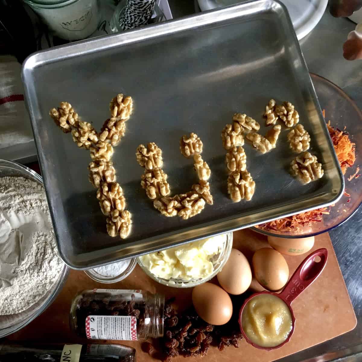 Purple carrot cake ingredients on a cutting board with toasted walnuts spelling YUM on a small pan showing the best carrot cake recipe ingredients with walnuts.