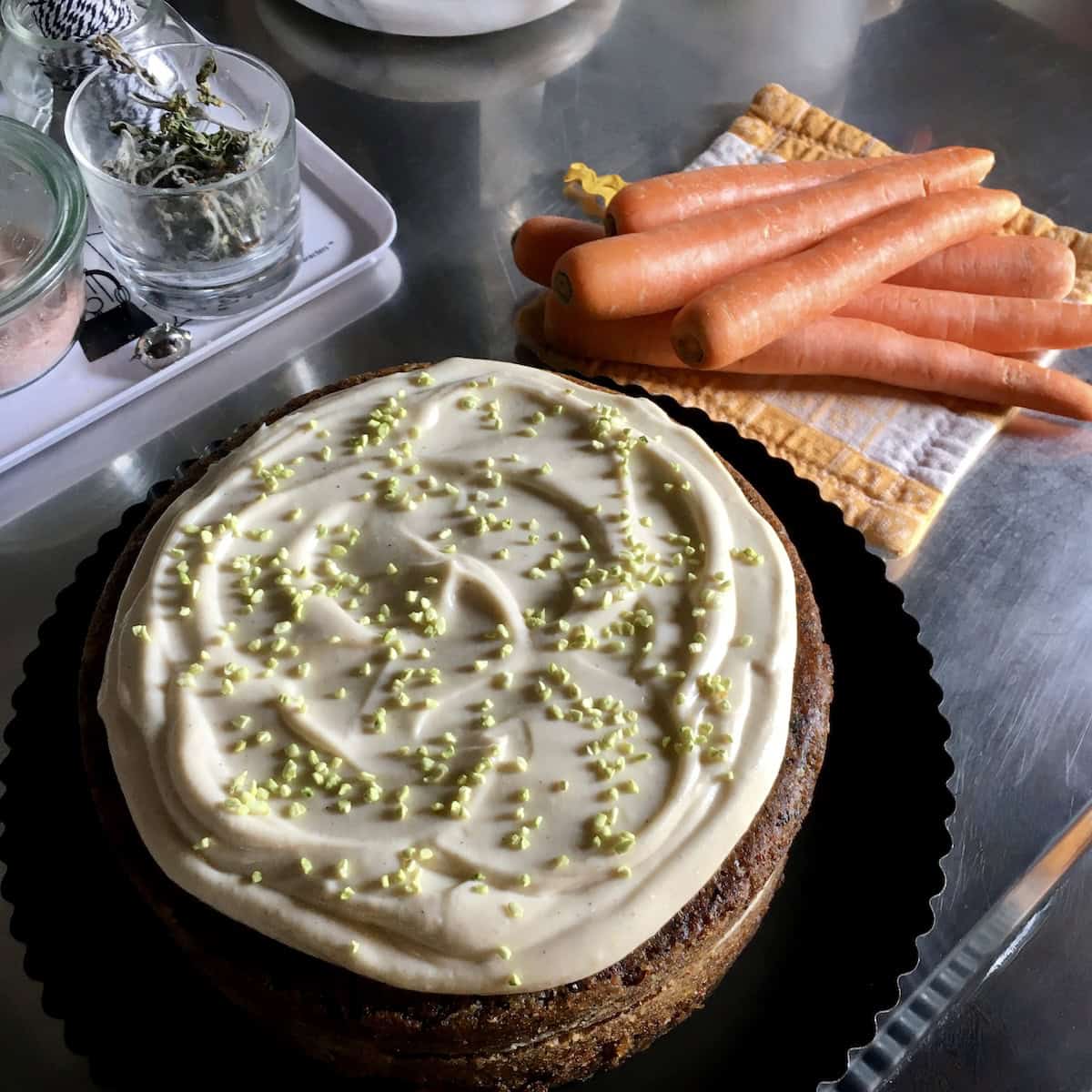 Double layer carrot cake with vanilla bean cream cheese frosting and pistachio sprinkles on a cake platter, whole carrots in the background.