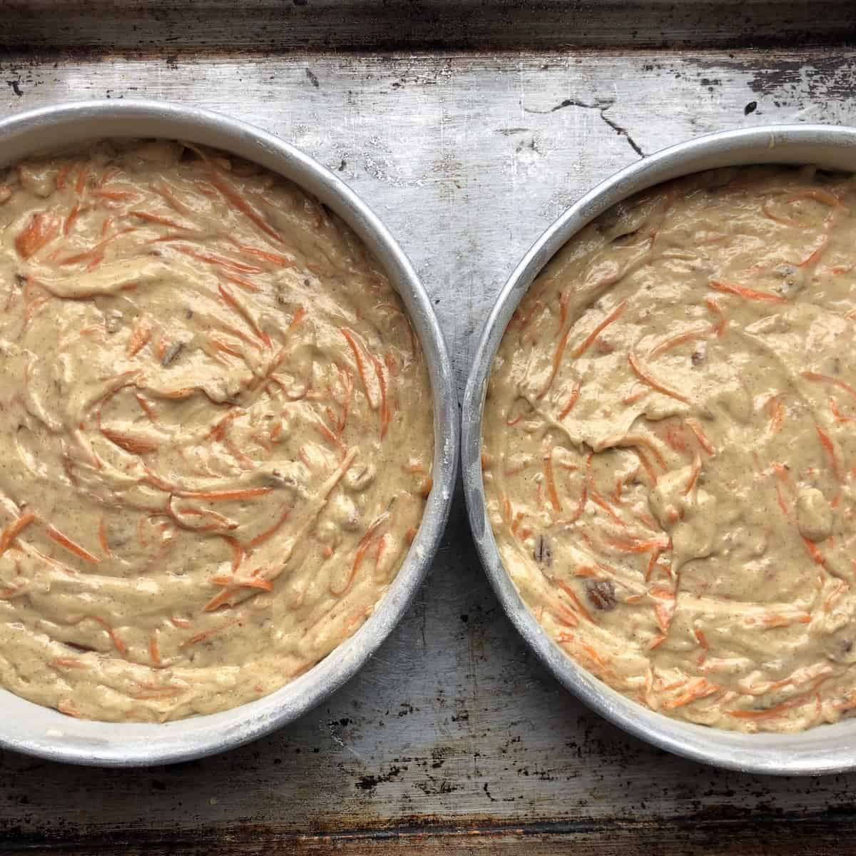 Orange carrot cake batter with toasted pecans and no raisins poured into two prepped 8-inch cake pans, slightly lighter in color than the purple version.