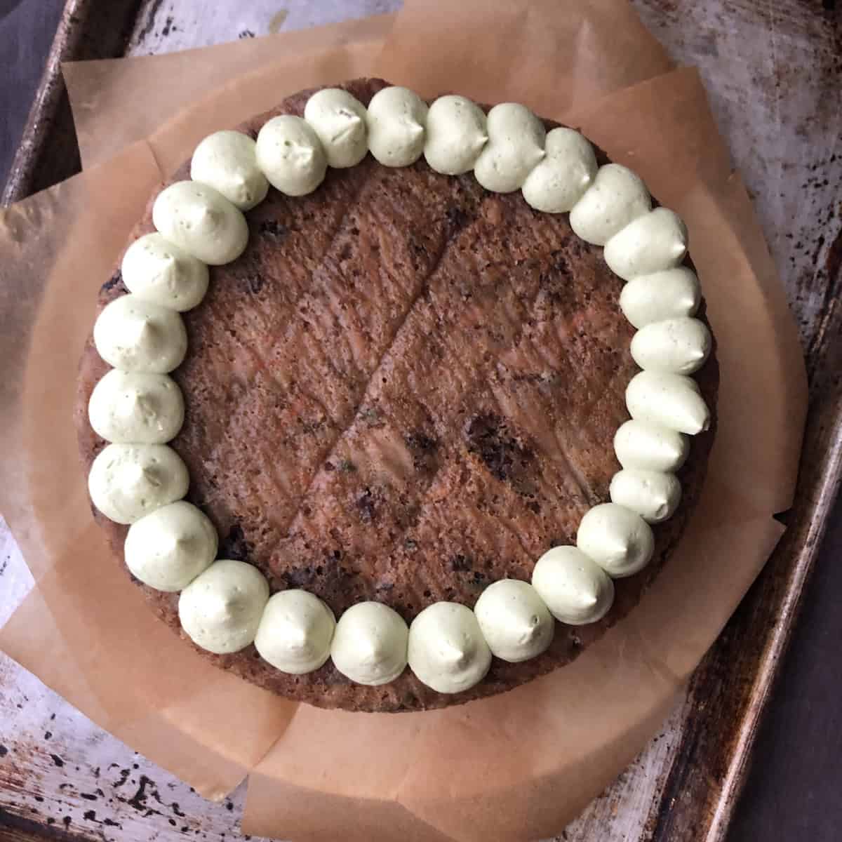 A piping bag adding kiss-shaped dollops of matcha cream cheese mascarpone frosting across the top of an orange carrot cake with pecans, without raisins, working from the outer edge inward to cover the whole surface.