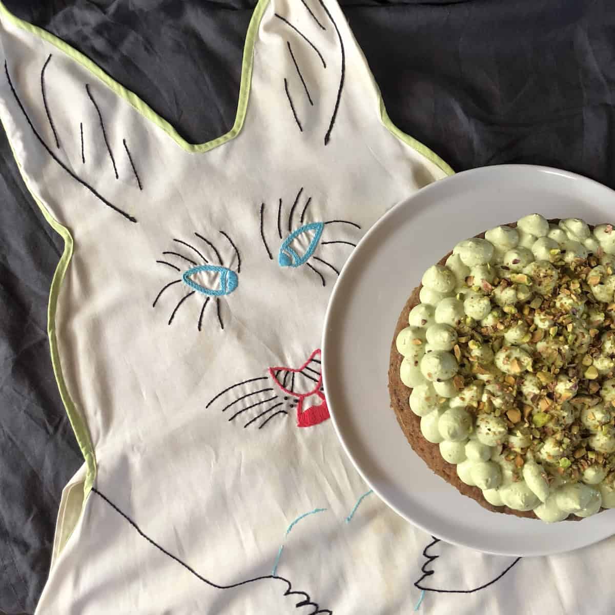 A vintage hand-stitched Easter bunny apron lying next to an orange carrot cake with matcha cream cheese mascarpone frosting and toasted pecans, the perfect Easter carrot cake centerpiece.