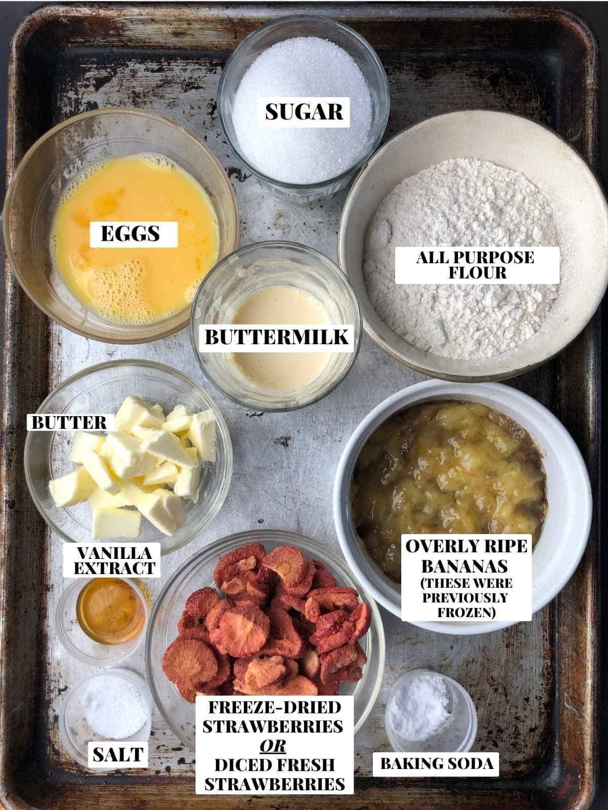 Labeled strawberry banana bread ingredients on a sheet pan including flour, sugar, butter, vanilla extract, mashed bananas, salt, baking soda, freeze-dried strawberries, and buttermilk substitute.
