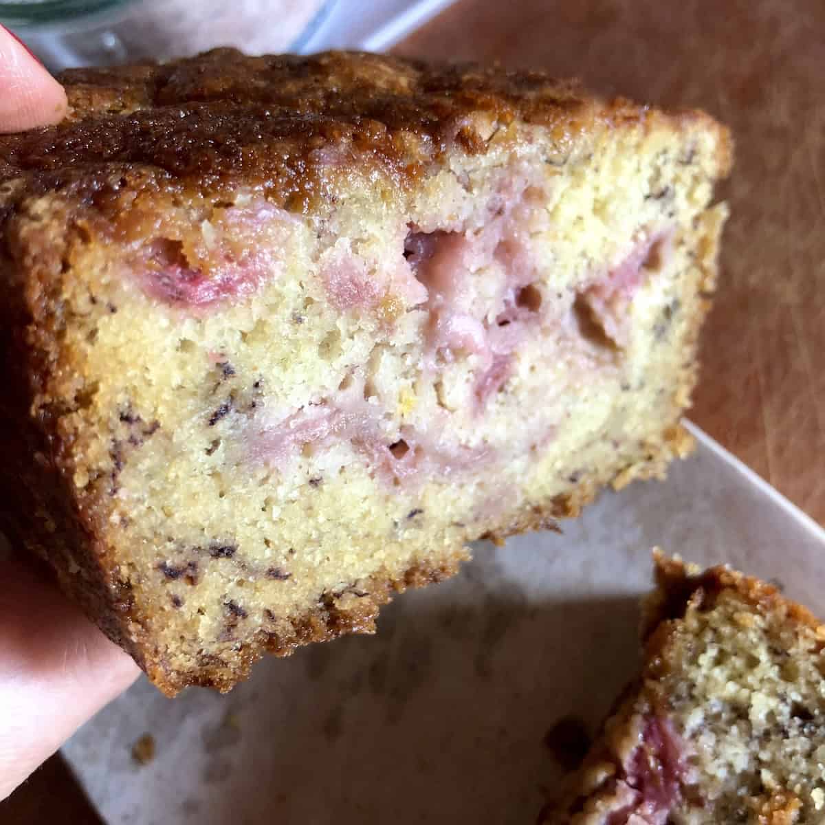 Hand holding a slice of strawberry banana bread showing the moist crumb studded with pink fresh strawberry pieces.
