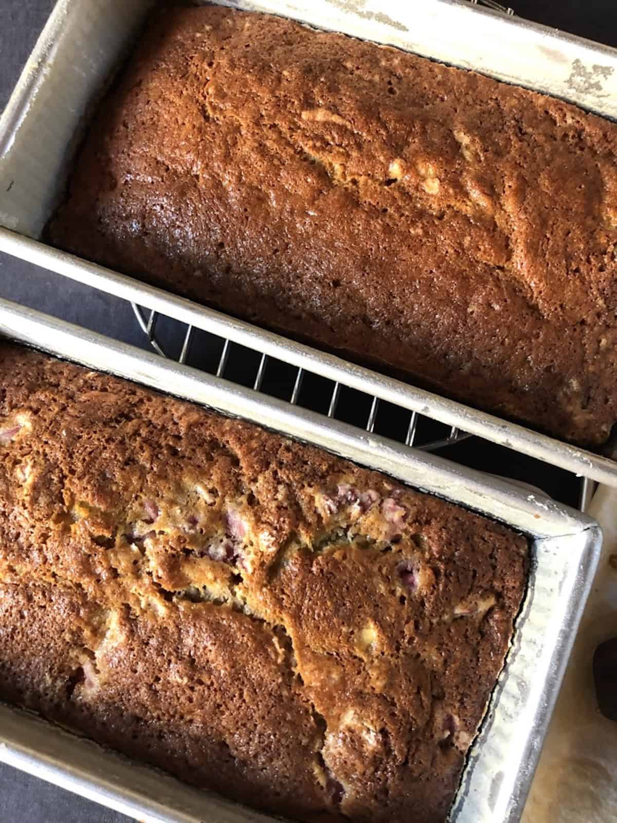 Two baked strawberry banana bread loaves cooling in their pans next to overripe bananas on parchment paper.
