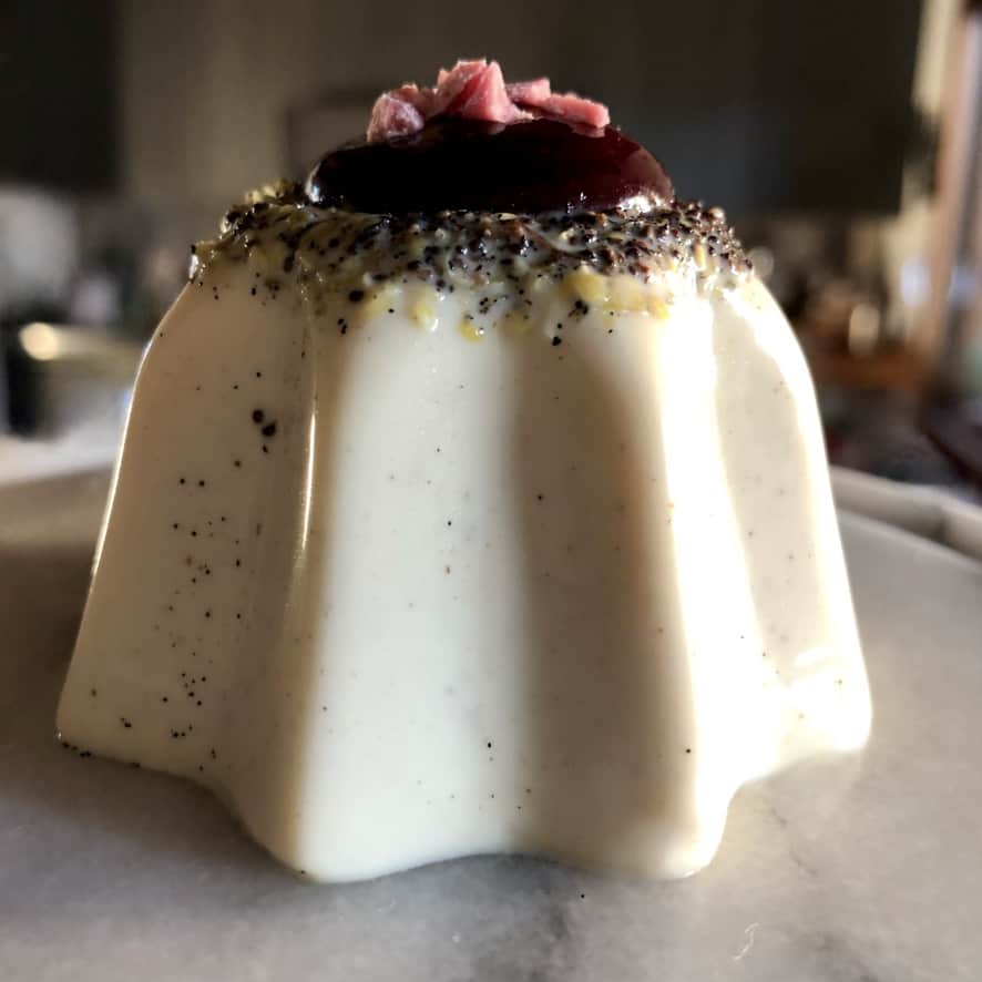 vanilla bean-speckled white chocolate lemon poppyseed panna cotta in the shape of a Pandora star with the seeds and lemon zest visibly concentrated on top with a perfect squeeze of dark berry sauce in the middle and chopped pink white chocolate pieces as decoration and a spoonful removed