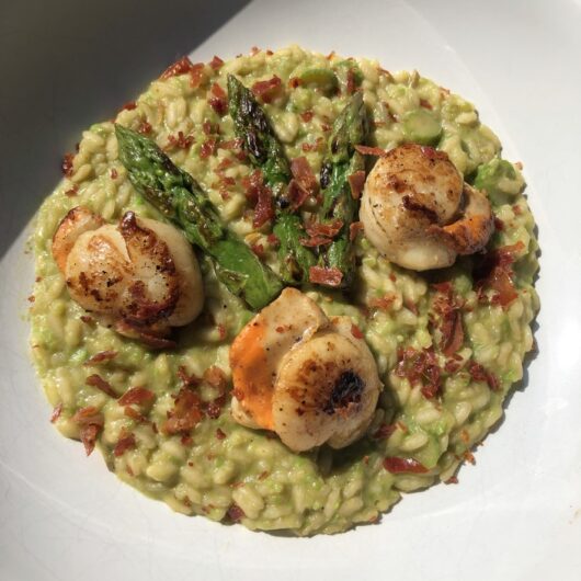 a pasta bowl plate filled with spring green asparagus risotto with three pan seared spears and three pan seared scallops on top with a sprinkling of crumbled crispy-fried prosciutto on top
