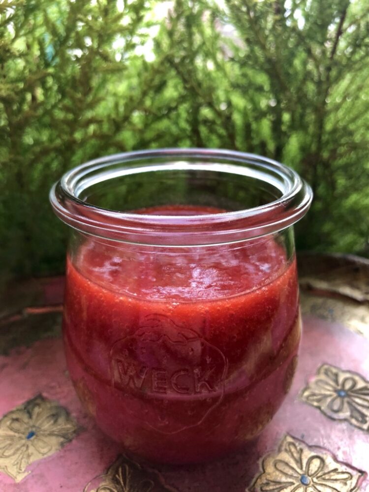 a glass Weck tulip jar filled with strawberry blood orange sauce on a pink and gold vintage venetian wooden tray with the lime green mini cypress trees behind it