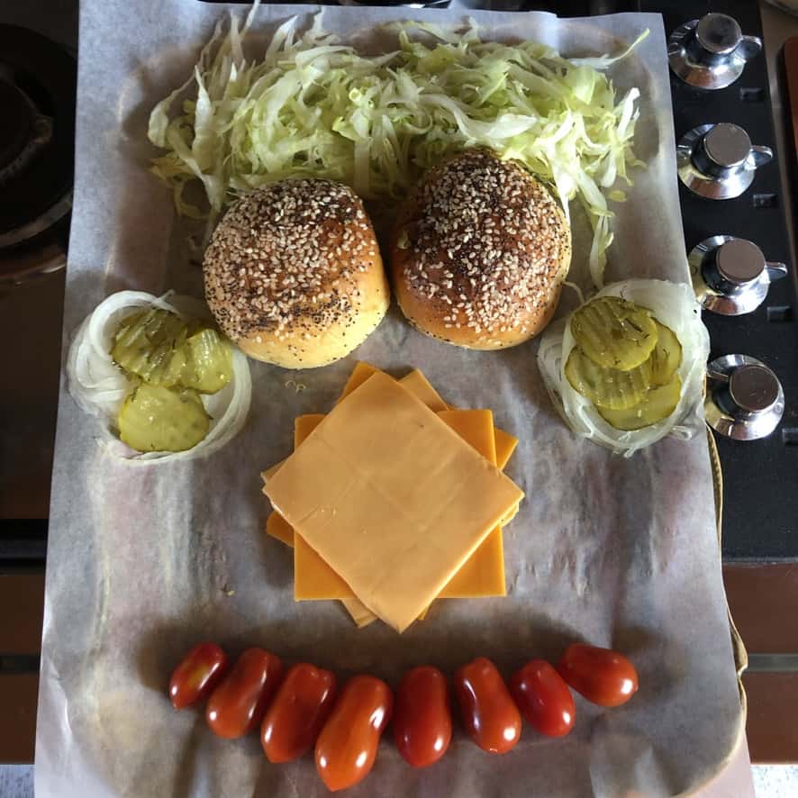 a happy face made out of all of the cheeseburger ingredients including the buns for eyes, shredded lettuce for hair, cheese for the nose, grape tomatoes for the mouth, and onions with pickles stacked on top for the ears