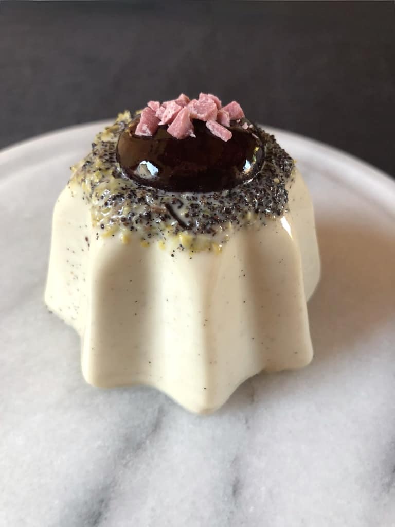 vanilla bean-speckled white chocolate lemon poppyseed panna cotta in the shape of a Pandora star with the seeds and lemon zest visibly concentrated on top with a perfect squeeze of dark berry sauce in the middle and chopped pink white chocolate pieces as decoration and a spoonful removed