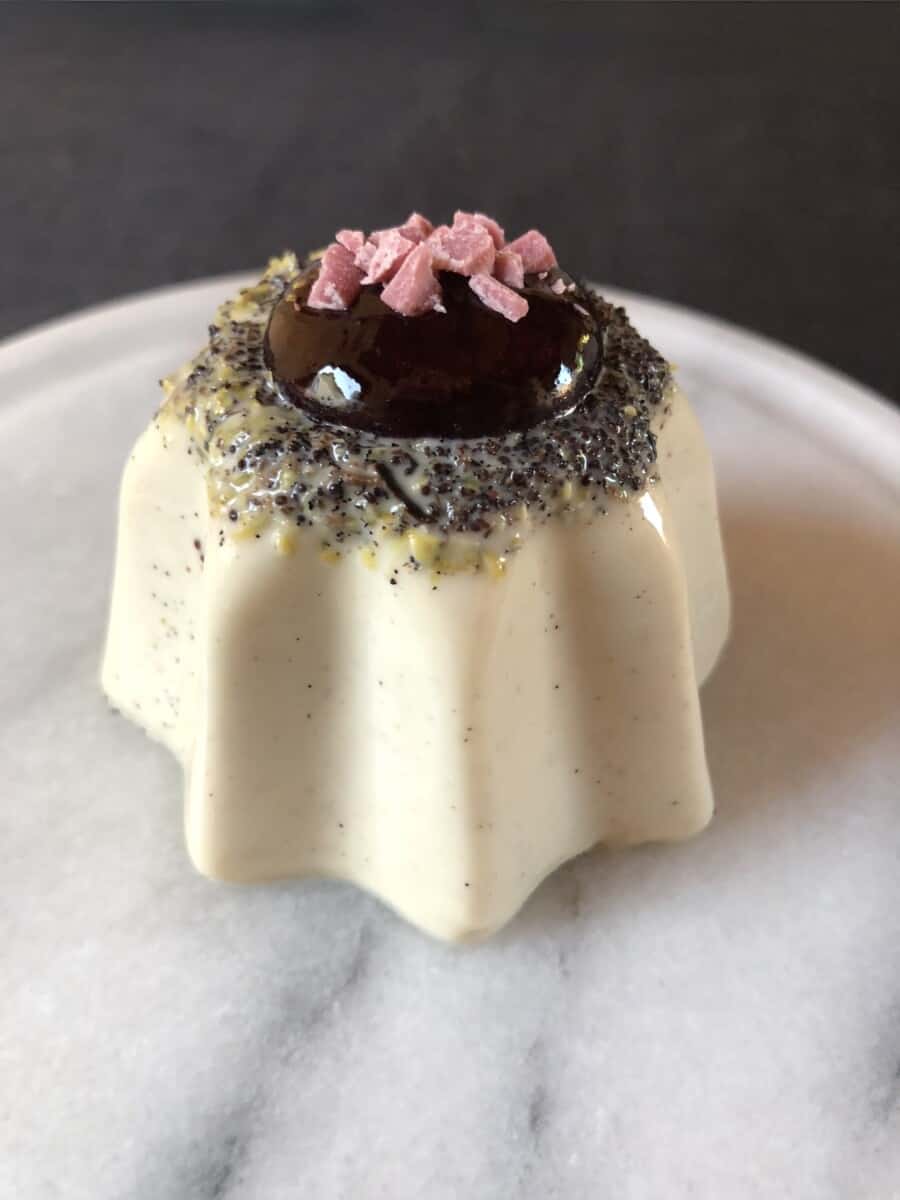 vanilla bean-speckled white chocolate lemon poppyseed panna cotta in the shape of a Pandora star with the seeds and lemon zest visibly concentrated on top with a perfect squeeze of dark berry sauce in the middle and chopped pink white chocolate pieces as decoration
