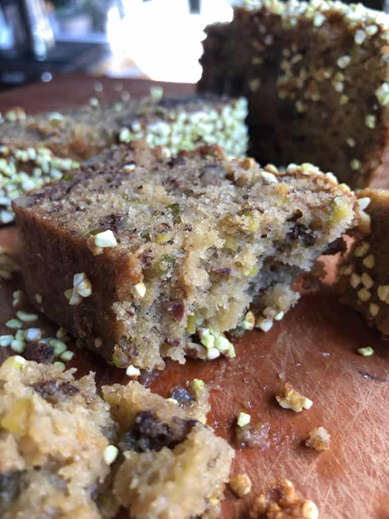 A slice of moist banana nut bread with freeze-dried pistachios on top and nuts throughout the crumb on a cutting board.
