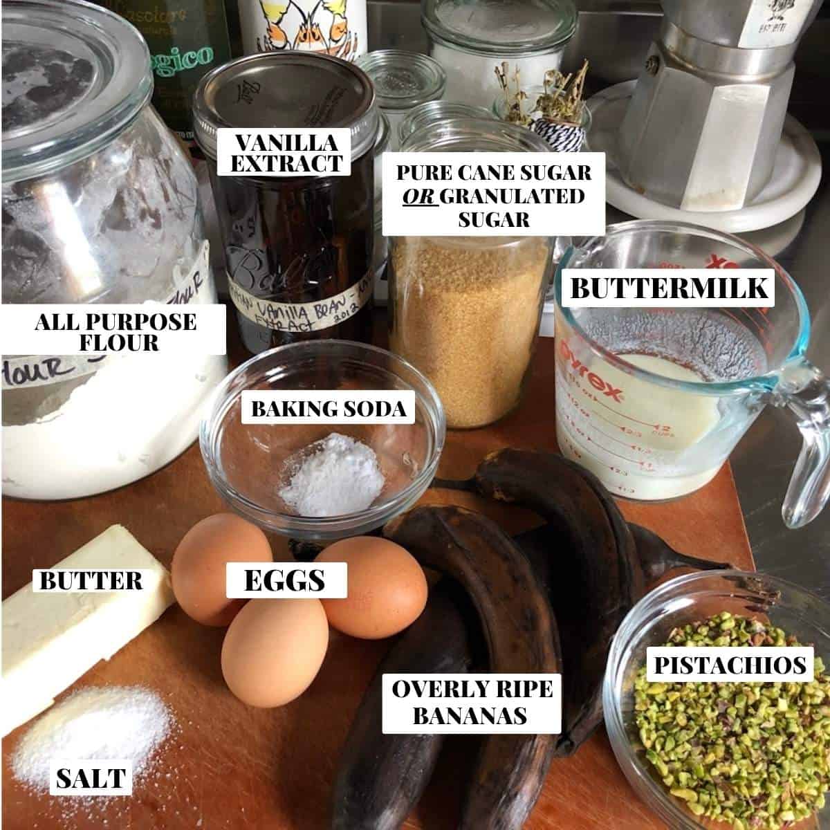 Labeled pistachio banana bread ingredients on a countertop including ripe bananas, flour, baking soda, butter, eggs, chopped pistachios, buttermilk, sugar, salt, and vanilla extract.
