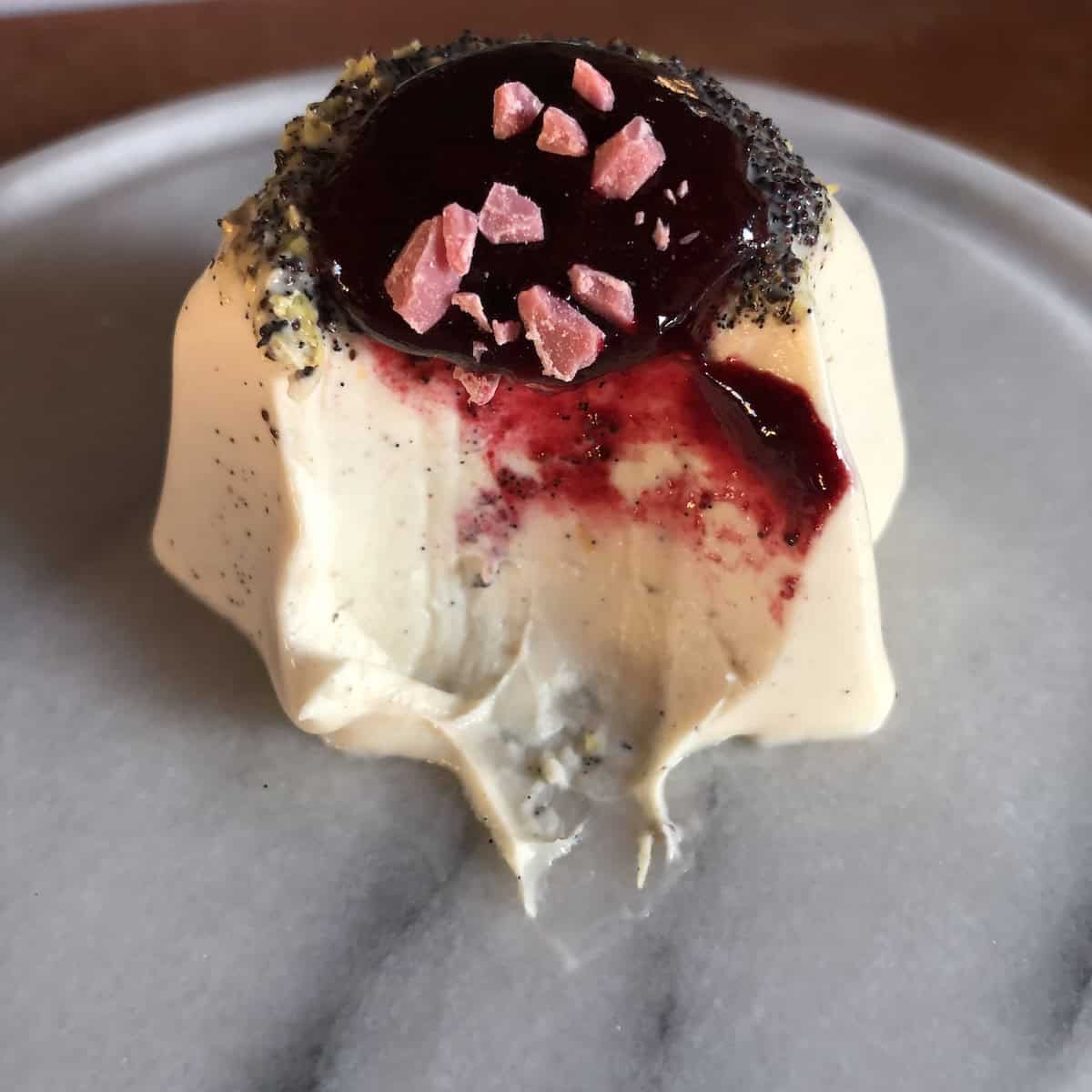 Homemade vanilla bean panna cotta with visible vanilla bean seeds topped with wild berry coulis sauce and a bite removed showing the perfect creamy jiggly texture, a perfect Italian Easter dessert recipe.