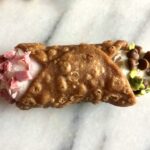 a beautiful golden brown cannoli with pink strawberry ricotta cream and pink white chocolate sprinkleeson one end and pale pistachio green ricotta cream on the other end with mini chocolate chips and pistachios