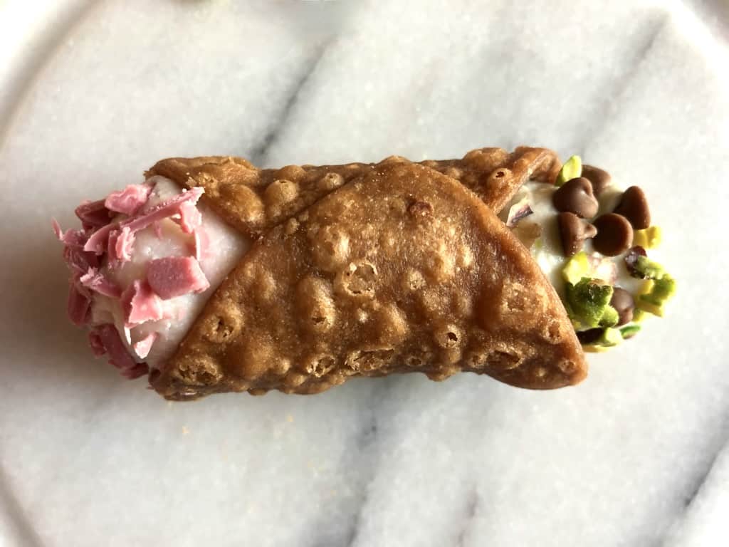 a beautiful golden brown cannoli with pink strawberry ricotta cream and pink white chocolate sprinkleeson one end and pale pistachio green ricotta cream on the other end with mini chocolate chips and pistachios