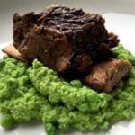 A single braised bone-in beef short rib sitting on top of a mound of bright green mushy peas side dish.