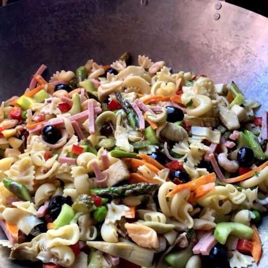 a 16 inch wok filled with a beautiful rainbow-colored pasta salad with a pink hibiscus barely visible in the background