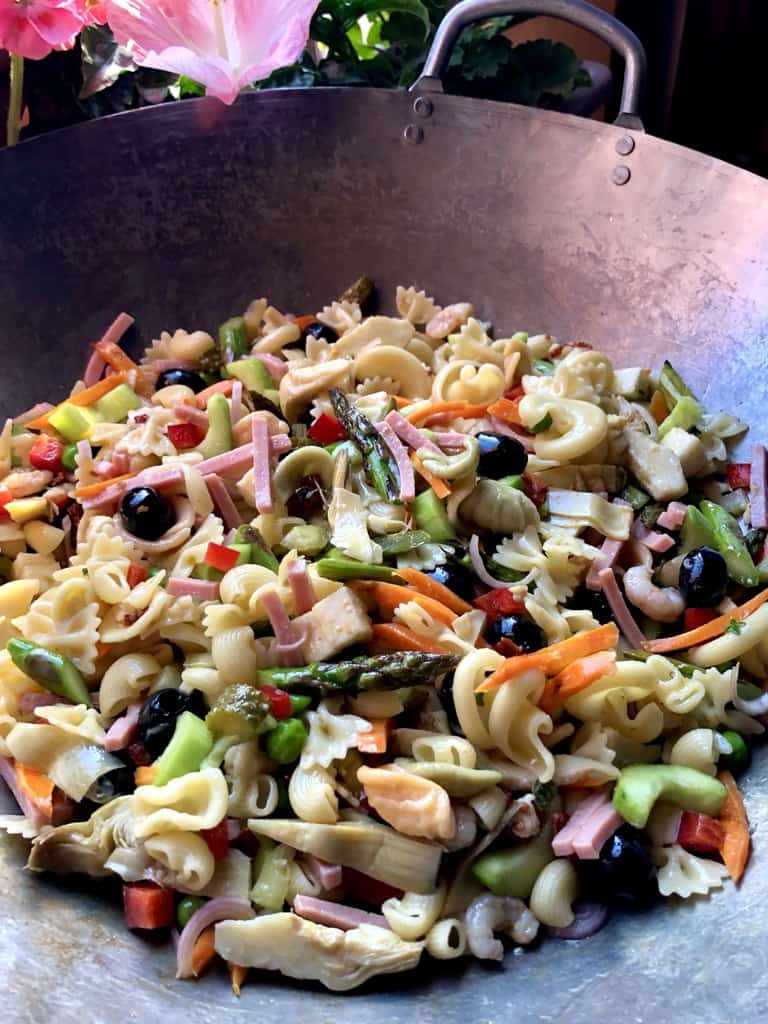 a 16 inch wok filled with a beautiful rainbow-colored pasta salad with a pink hibiscus barely visible in the background