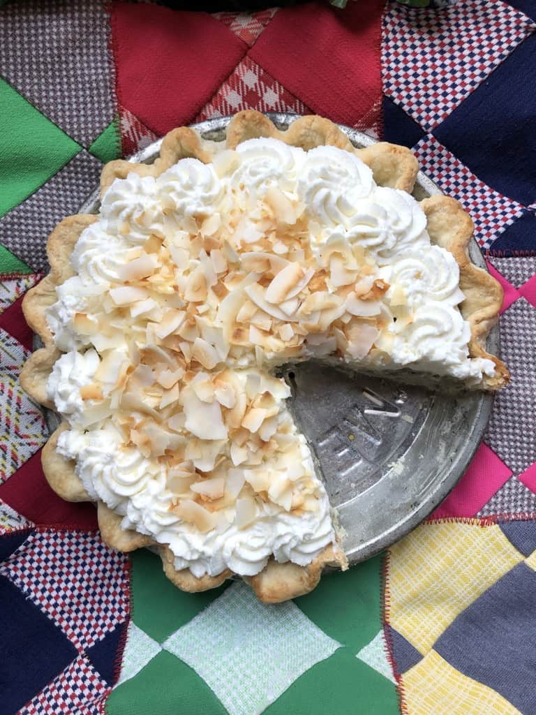 Coconut cream pie with black sesame on a patchwork quilt with one slice removed showing the grey coconut cream filling.