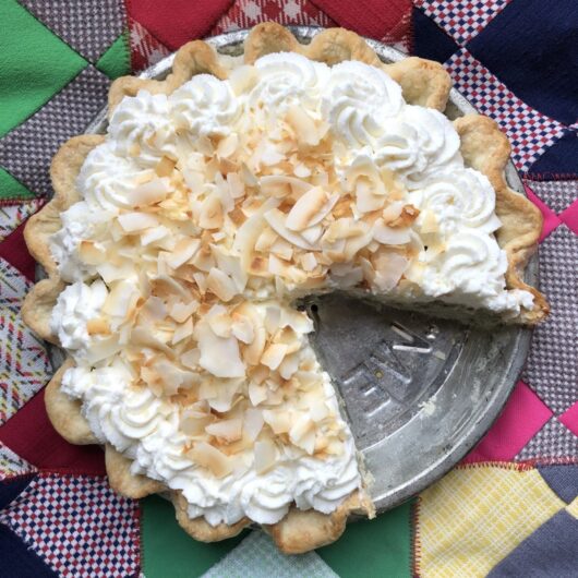 a beautiful coconut cream pie with whipped cream and toasted large flake coconut piled high with 2 slices removed to reveal the vintage pie tin