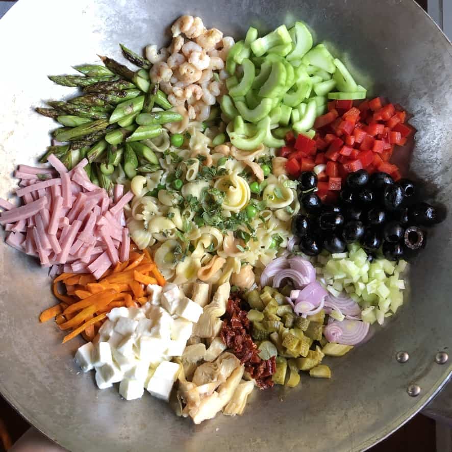 Mediterranean pasta salad ingredients (raw and cooked and cured items) all bundled together in singular mounds in a wok before being stirred together.