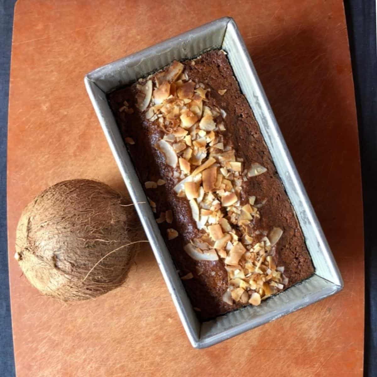 A fully baked banana bread with coconut and chocolate chips in a USA Pan loaf pan next to a whole coconut on a cutting board, banana coconut chocolate chip bread recipe.