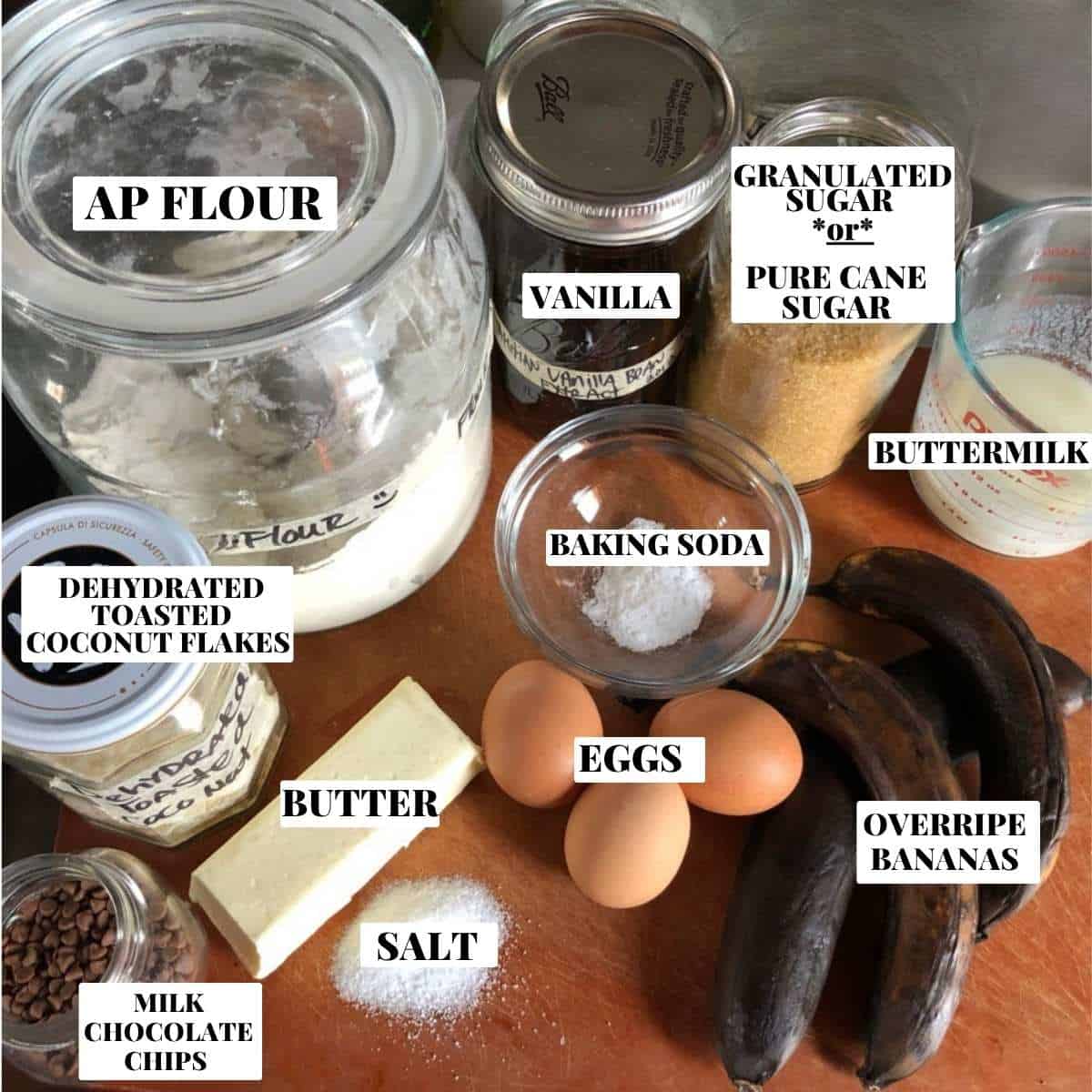 Labeled banana coconut chocolate chip bread ingredients on a cutting board including ripe bananas, shredded coconut, milk chocolate chips, flour, sugar, eggs, vanilla, dehydrated coconut flakes, buttermilk, baking soda, salt, and butter for a banana bread with coconut and chocolate chips.