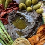 Black chickpea hummus in a serving platter (closeup shot) with a well in the middle filled with EVOO and surrounded by sauteed carrot slices, grilled asparagus, seared onion, sundried tomatoes, cornichon pickles, and large green olives with pits.