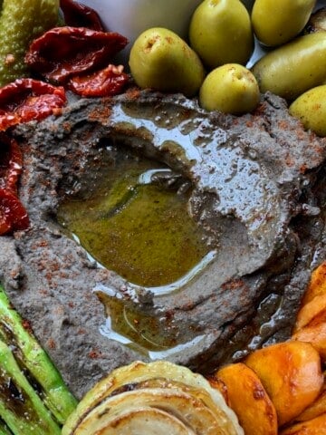 Black chickpea hummus in a serving platter (closeup shot) with a well in the middle filled with EVOO and surrounded by sauteed carrot slices, grilled asparagus, seared onion, sundried tomatoes, cornichon pickles, and large green olives with pits.