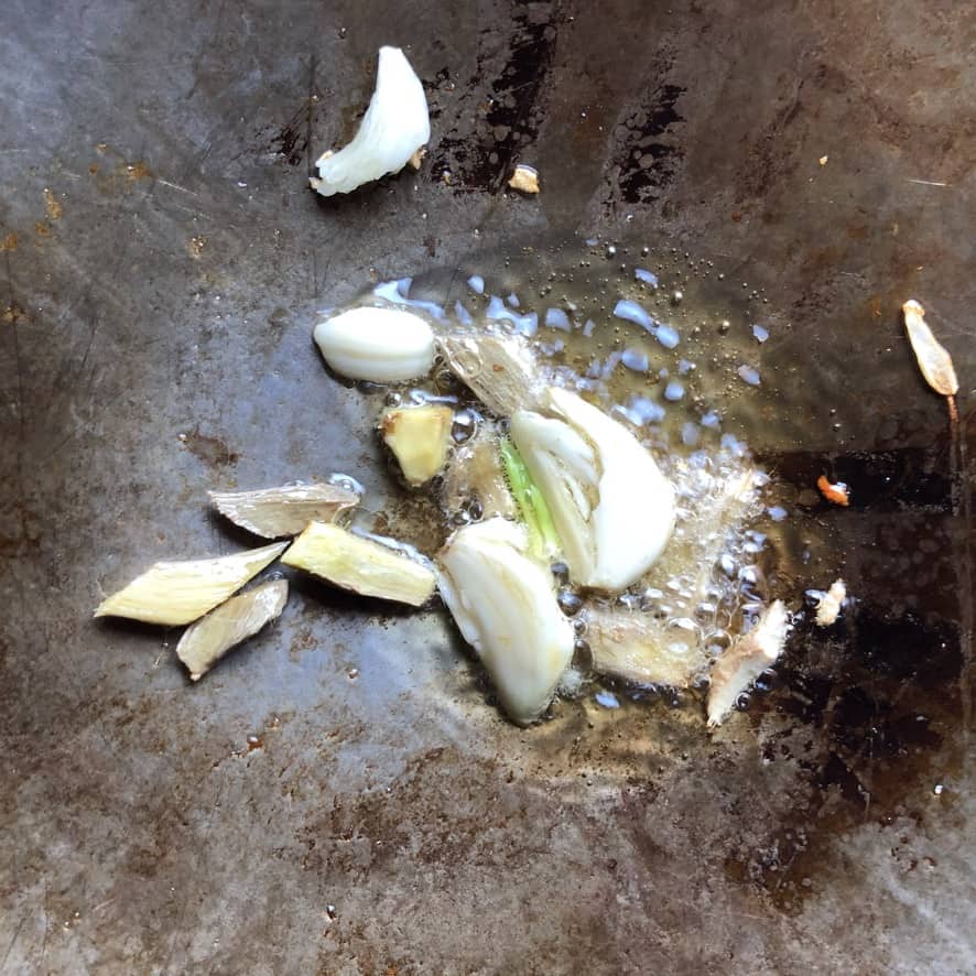 smashed garlic cloves and sliced ginger cooking in hot oil inside of a wok