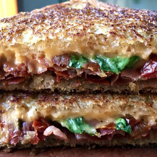 closeup of crispy prosciutto and sun dried tomatoes with melty cheese and speckles of green arugula inside a just-cooked grilled cheese sandwich