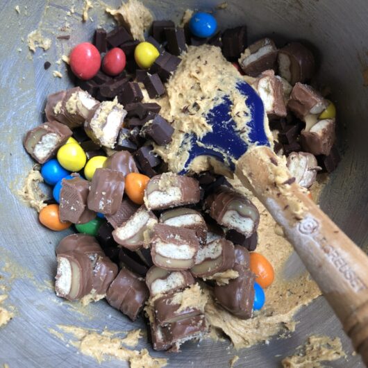 chocolate chip cookie dough with a pile of peanut M&M's, roughly chopped twix bars, Lindt dark chocolate chunks and pastel colored Italian Smarty candies before being mixed in