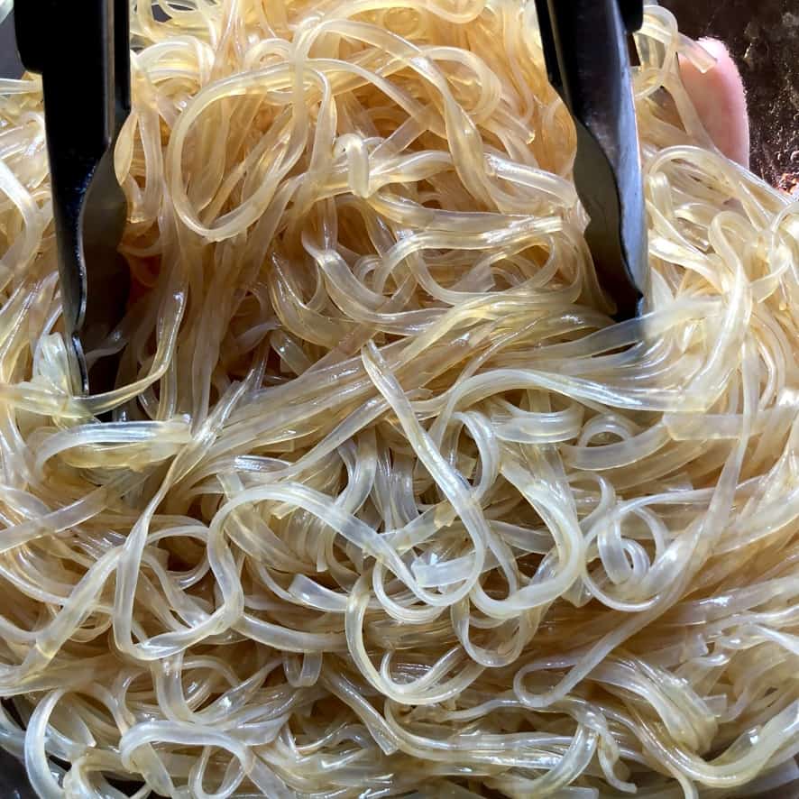 cooked glass noodles with some dark soy sauce and a splash of oil added to them and tossed making them a light brown sugar color and a pair of tongs resting in the bowl