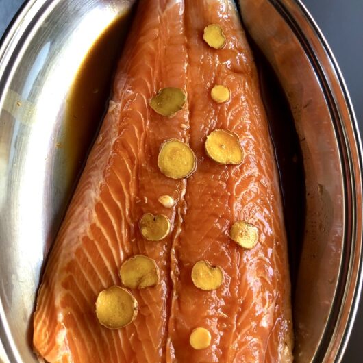 a whole side of salmon in the marinade with sliced ginger rounds resting on top of it's orange flesh