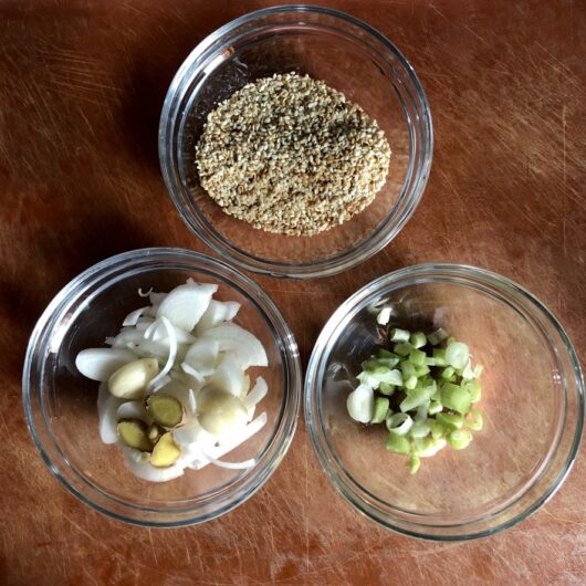 a trio of small glass prep bowls with one containing 2 smashed garlic cloves, one with sliced scallions, and the last with toasted sesame seeds