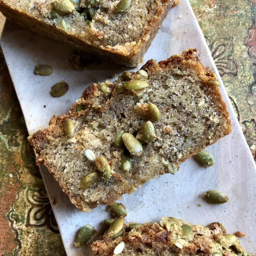 sliced pumpkin spice banana bread on a pink rectangular marble serving tray with toasted crunchy sugary pumpkin seeds coating the tops of each slice