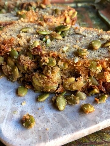 A slice of banana bread with pumpkin seeds on a pink marble tray showing the moist crumb and crunchy seed topping.
