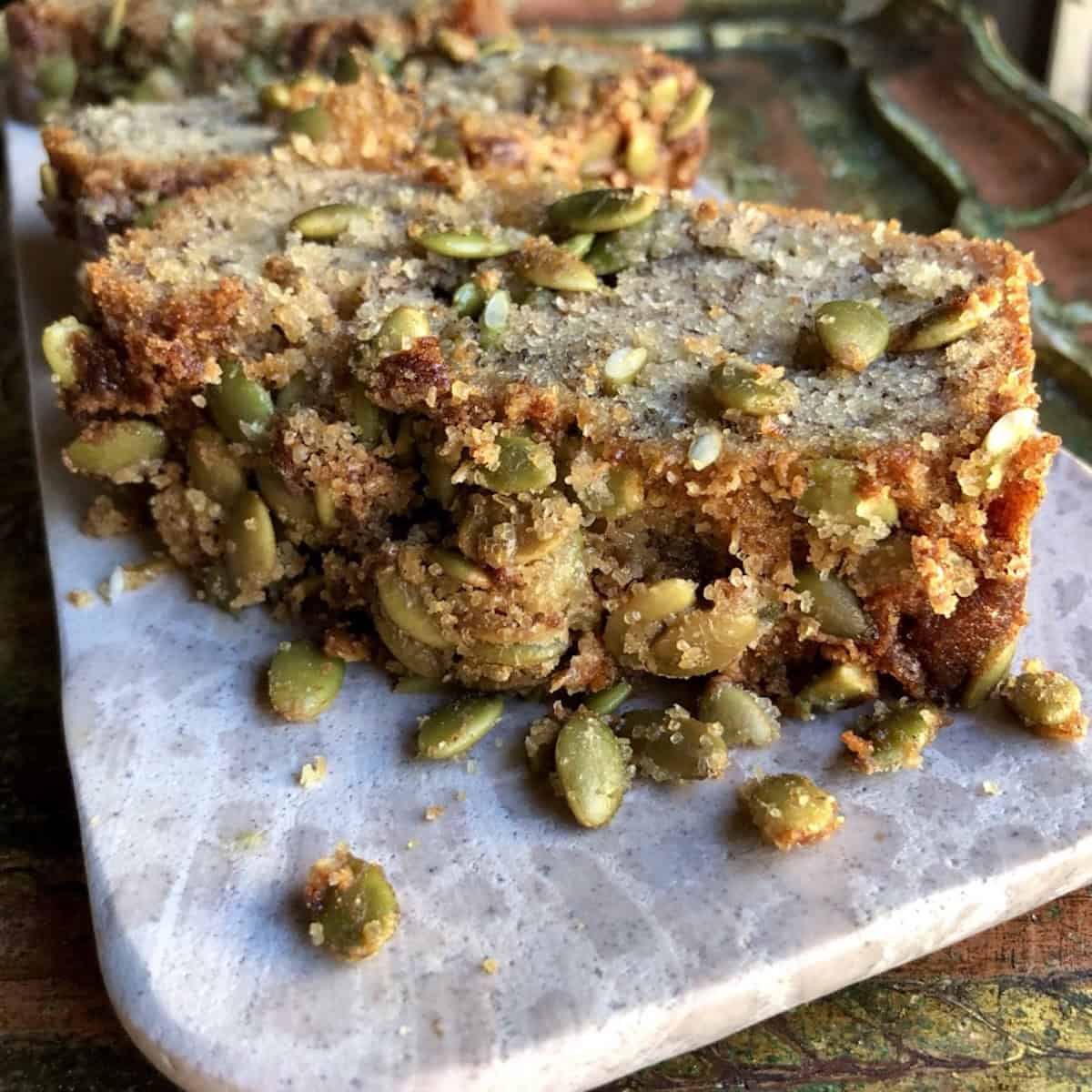 A slice of banana bread with pumpkin seeds on a pink marble tray showing the moist crumb and crunchy seed topping.