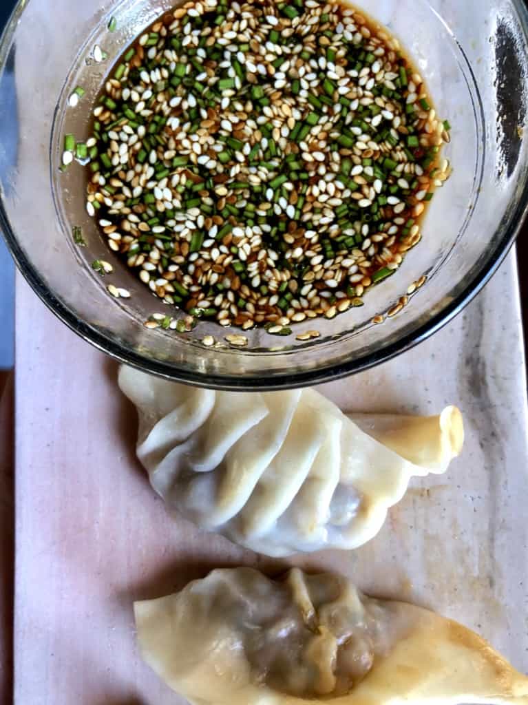 freshly steamed chicken dumplings on a marble serving tray with a top down view of the homemade dipping sauce with lots of chives and sesame seeds floating on top.