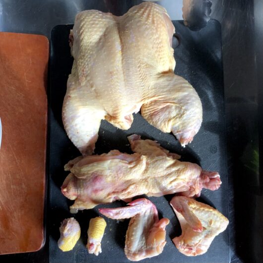 a whole chicken on a cutting board with the back, wings and tips of the legs removed and ready to be used in the stock