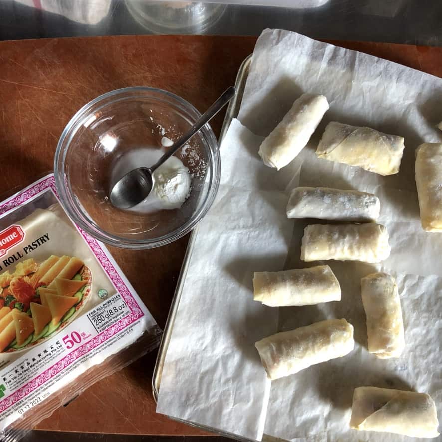 spring roll wrappers still in the package lying next to a bowl with a cornstarch slurry and just-filled and rolled spring rolls