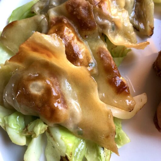 a layer of stacked golden brown and glossy pork and chive potstickers plated on top of sauteed cabbage with crispy spring rolls in the background