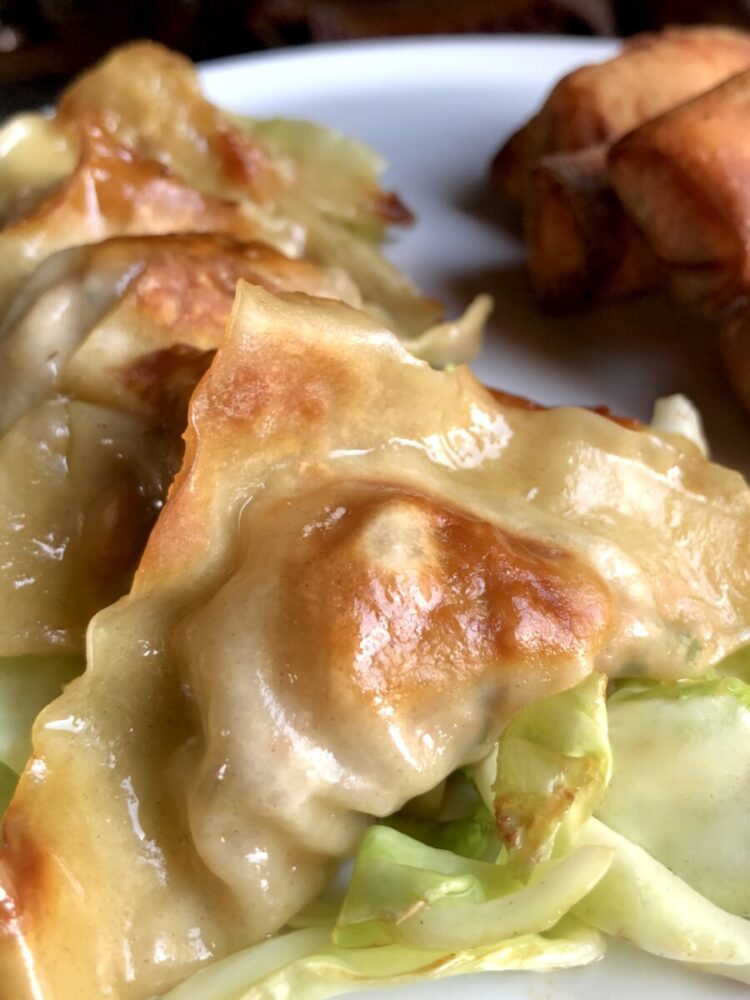 a layer of stacked golden brown and glossy pork and chive potstickers plated on top of sauteed cabbage with crispy spring rolls in the background