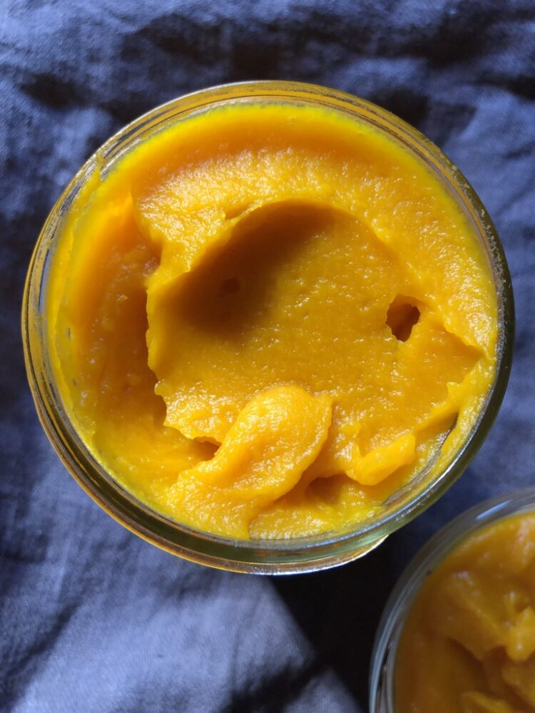 top down view of widemouth mason jar filled with bright orange homemade pumpkin purée sitting on top of a dark grey linen apron