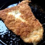 golden brown breaded turkey breast cutlet frying in a cast iron skillet with very little extra virgin olive oil