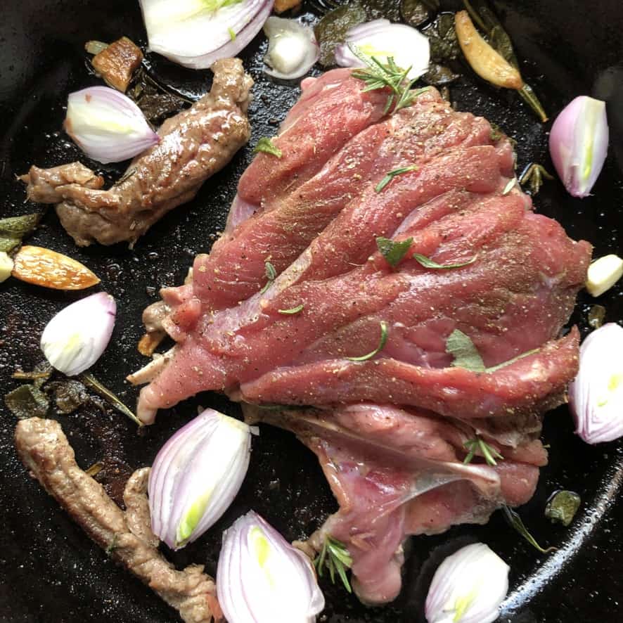 butterflied turkey leg lying in a skillet surrounded by shallots, herbs, and garlic cloves (meat side up)