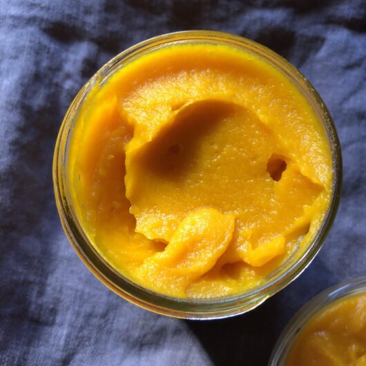 top down view of widemouth mason jar filled with bright orange homemade pumpkin purée sitting on top of a dark grey linen apron