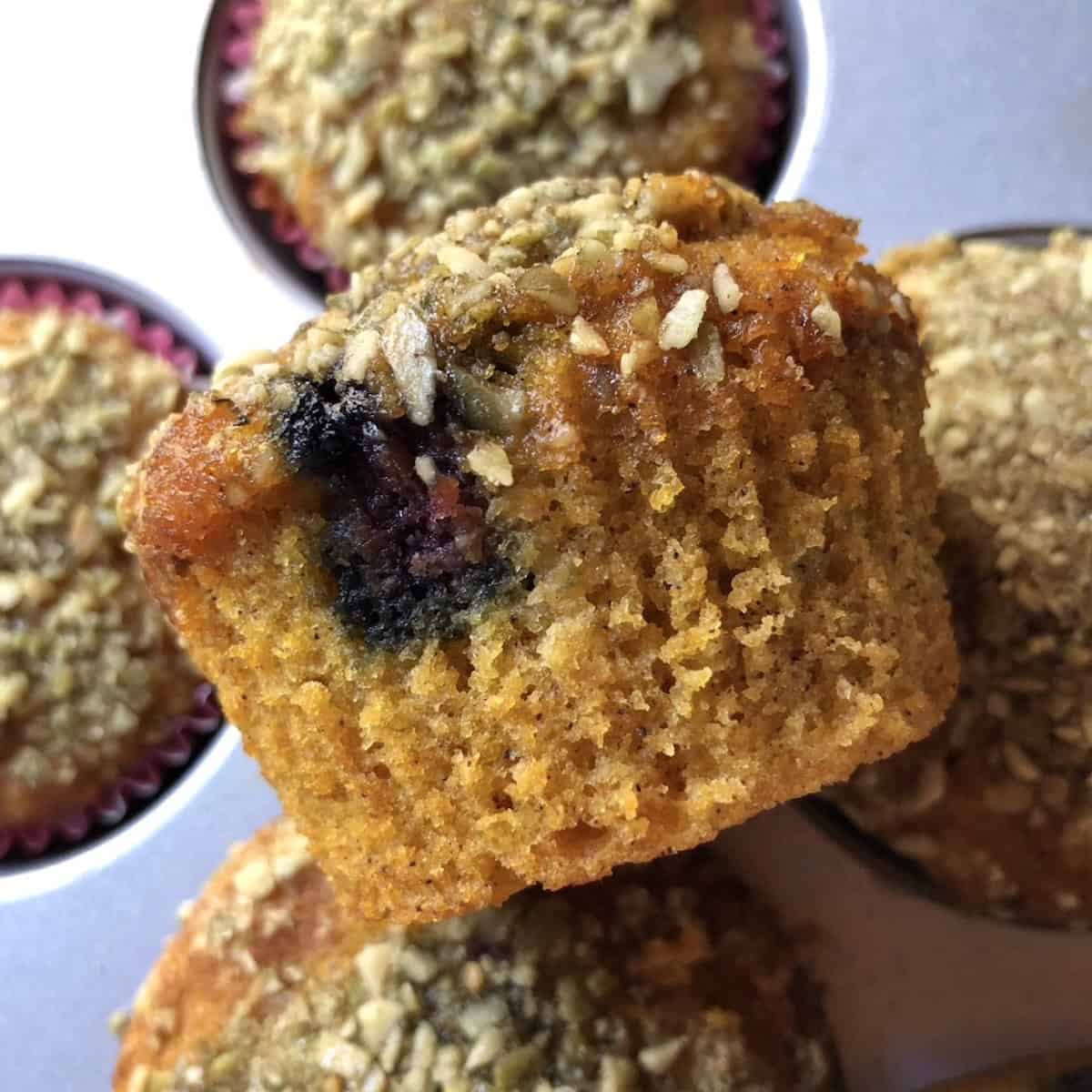 A blueberry pumpkin muffin with pumpkin seed crunch topping unwrapped from the liner showing berries in the tender crumb from the side.
