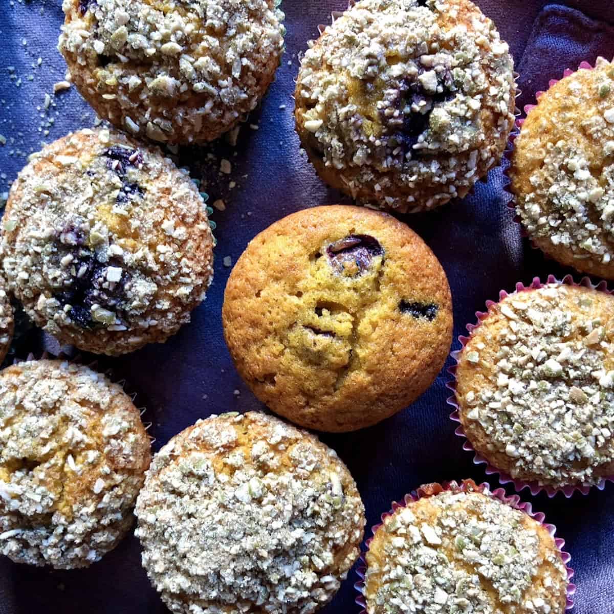 Tender golden brown blueberry pumpkin muffins with blueberries visible throughout the top with a single muffin in the center without any topping and the rest with pumpkin seed crunch topping to make it more bakery-style.
