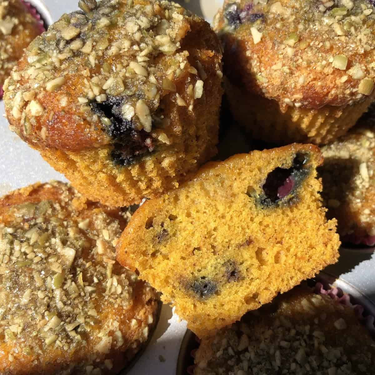 A blueberry pumpkin muffin sliced in half showing the moist tender crumb studded with blueberries with the rest of the muffins in the background.
